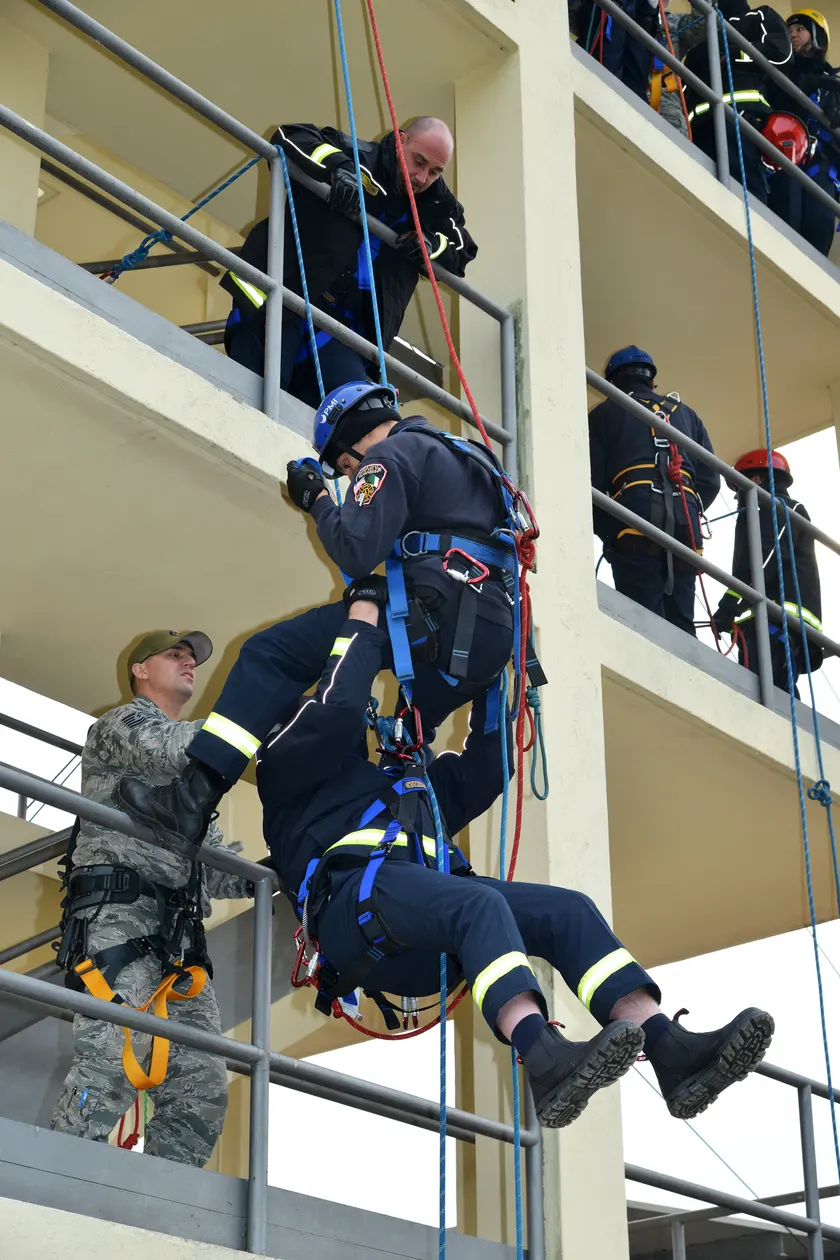Two firefighters in harnesses practice a rope rescue beside the open floors of a training tower